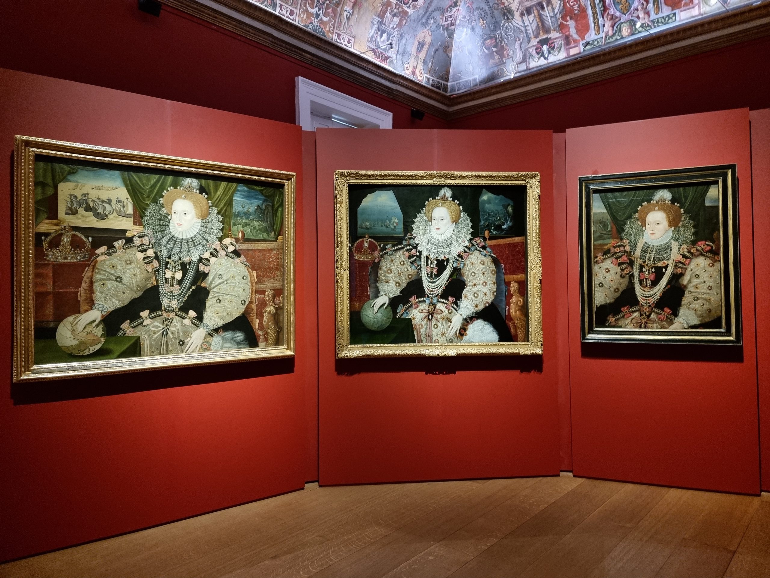 three portraits of Elizabeth I hung on red background. Elizabeth wears a lace ruff, and lots of pearsl, while her dress features bows. Her hand rests on a globe, with a crown next to her, and the Armada battle goes on in the background, seen through the windows