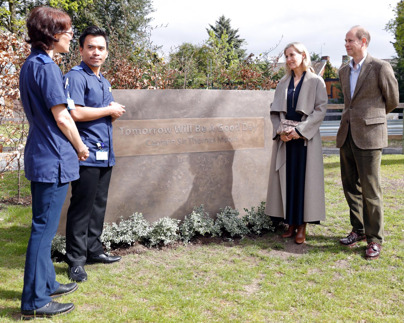 the earl and countess of wessex stand next to a stone plinth reading 'tomorrow will be a good day', the saying of captain tom moore. edward and sophie stand on the right, wearing a tweed jacket and grey long coat respectively, as they chat to two nurses in uniform