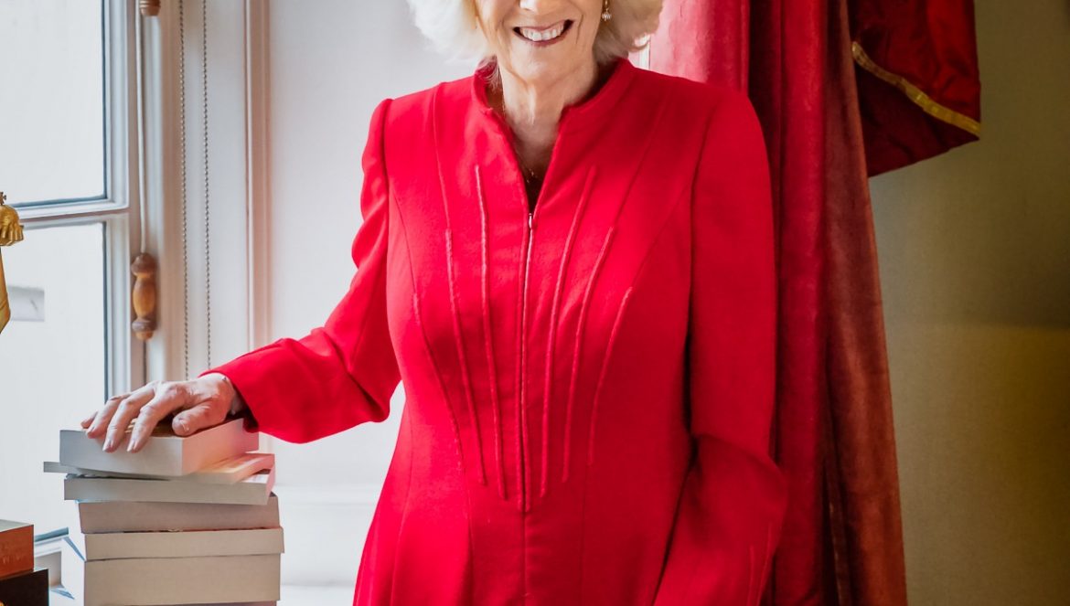 Camilla, The Duchess of Cornwall, in a red coat dress with her hand on a pile of books to launch her Reading Room