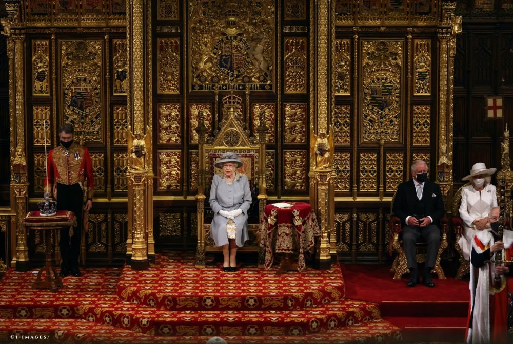 wide angle showing queen sitting on a raised throne in the house of lords, with a small side table with red velvet cloth next to her, that also holds her speech. she wears dove grey; prince charles and camilla sit on a lower part of the platform to her left.