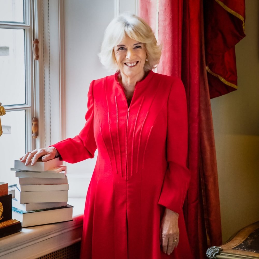 Camilla, The Duchess of Cornwall, in a red coat dress with her hand on a pile of books to launch her Reading Room